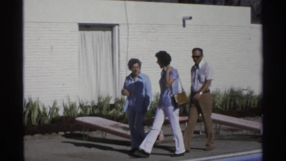 The adults continue past the pool, with the woman in blue leading and lounge chairs and white brick architecture behind them.