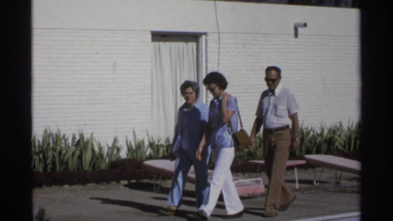 Three adults walk past a white motel wall, planted bed, and pink lounge chairs.
