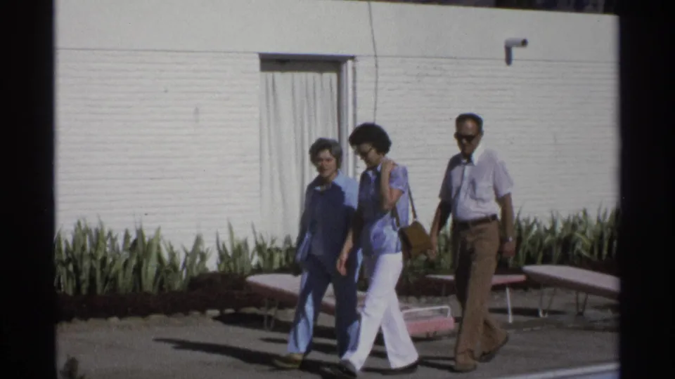 In Guaymas, Mexico, 1975, two women and a man walk outside a white motel with plants and pink lounge chairs nearby.