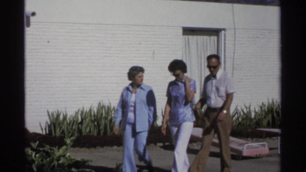 Two women and a man continue walking outside a white-walled Guaymas motel, with landscaping and lounge seating in view.
