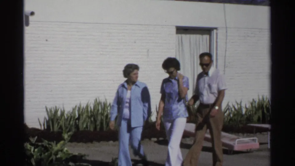 Two women and a man continue walking outside a white-walled Guaymas motel, with landscaping and lounge seating in view.