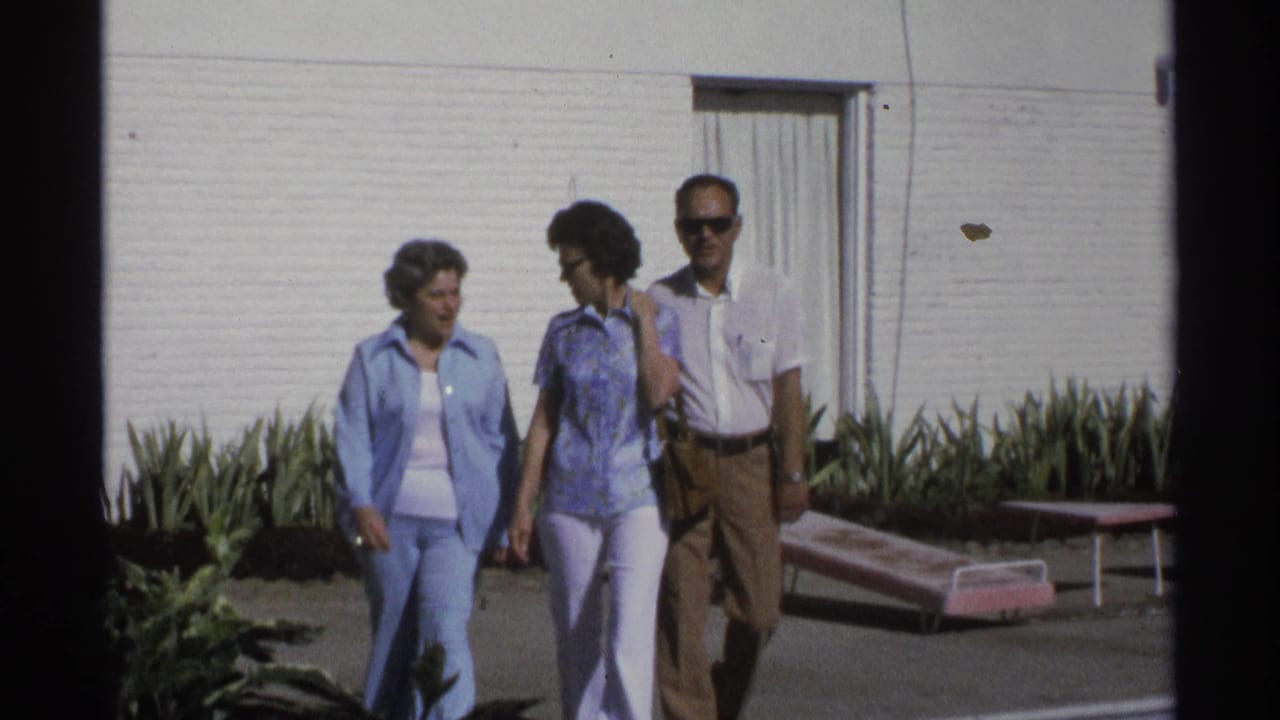 The walking group is shown closer outside the Guaymas motel, with 1970s clothing, sunglasses, plants, and lounge chairs visible.