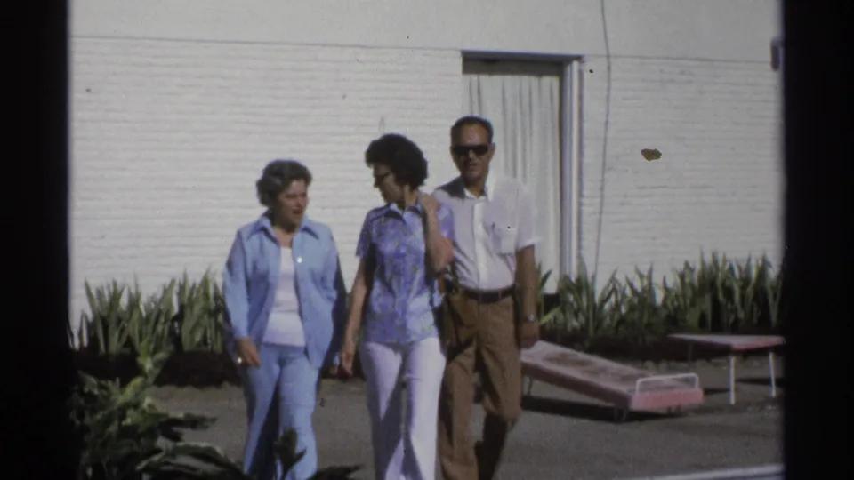 The walking group is shown closer outside the Guaymas motel, with 1970s clothing, sunglasses, plants, and lounge chairs visible.