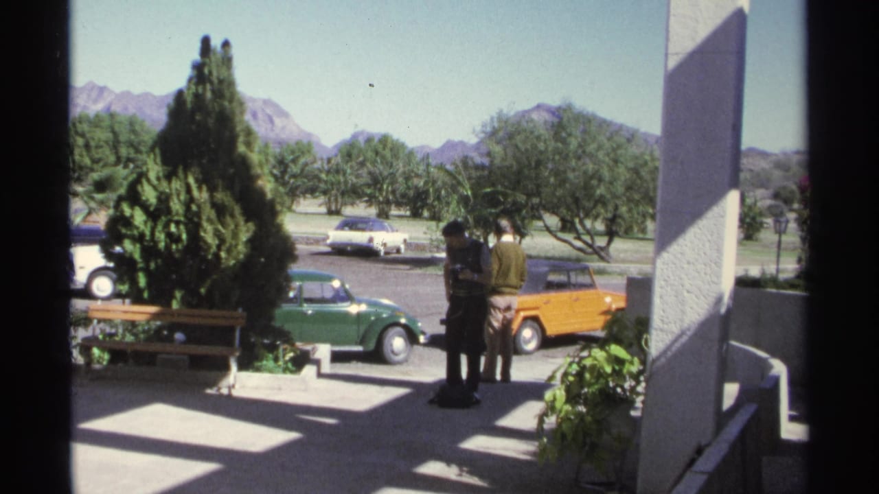 1975 Guaymas home-movie frame showing two adults by compact cars outside a shaded building, with trees and mountains in the distance.