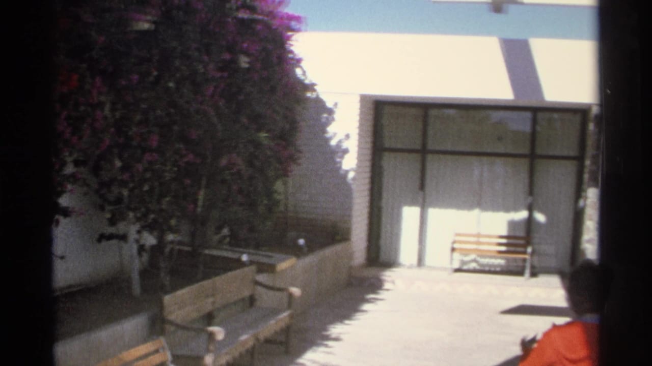 1975 Guaymas home-movie frame showing a bright courtyard with benches, glass doors, striped awning, purple blooms, and a woman entering the view.