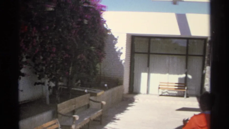 1975 Guaymas home-movie frame showing a bright courtyard with benches, glass doors, striped awning, purple blooms, and a woman entering the view.