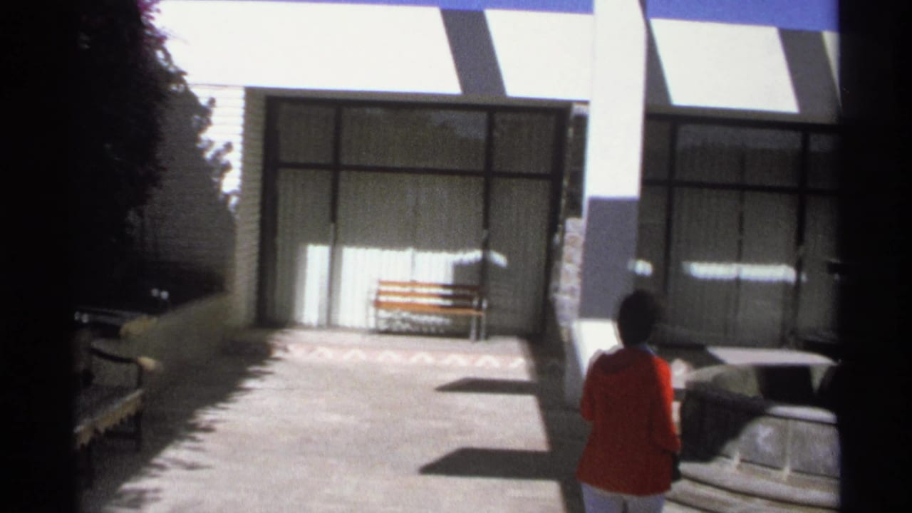 Woman in a red jacket walks across a bright courtyard toward benches and glass-fronted doors.