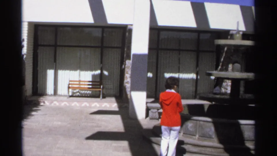 1975 Guaymas home-movie frame showing the woman from behind near a stone fountain, with courtyard benches and glass-fronted doors in sun.