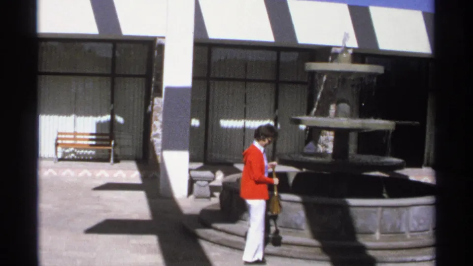 1975 Guaymas frame of a woman in a red jacket standing beside a tiered stone fountain in a bright courtyard with benches and glass doors.