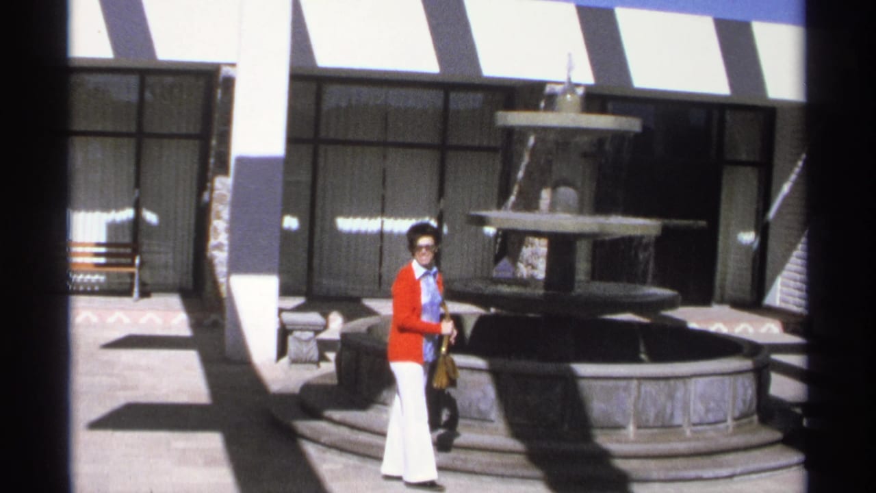 Woman in red cardigan and white pants standing beside a tiered courtyard fountain.