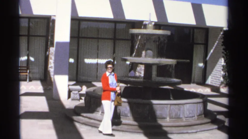 The woman remains near the courtyard fountain in Guaymas, with bright sunlight, striped awning, and building entrance visible.