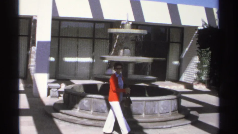 The woman walks through the Guaymas courtyard past the stone fountain, carrying a purse in bright 1970s travel footage.