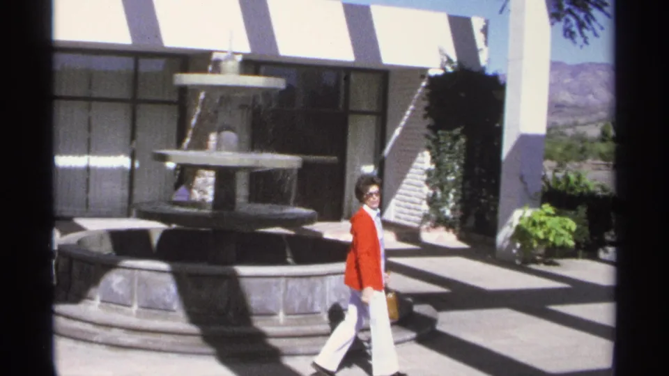 The woman leaves the fountain area in Guaymas, with stairs, plants, bright courtyard paving, and distant hills in view.