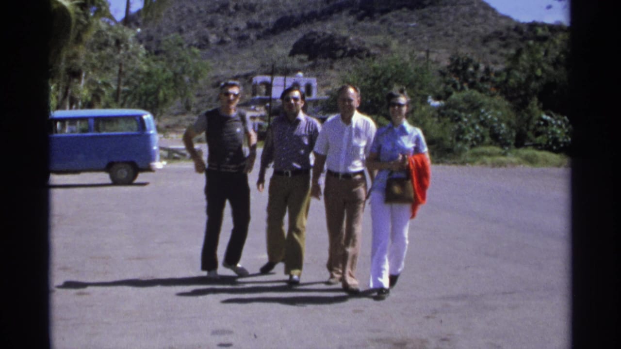 Four adults walk across a sunny paved area with a blue van, palms, and rocky hillside behind them.