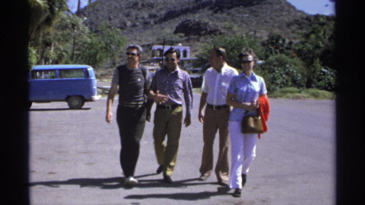 Four adults walk closer together across pavement, with a blue van and palm-lined hillside behind them.