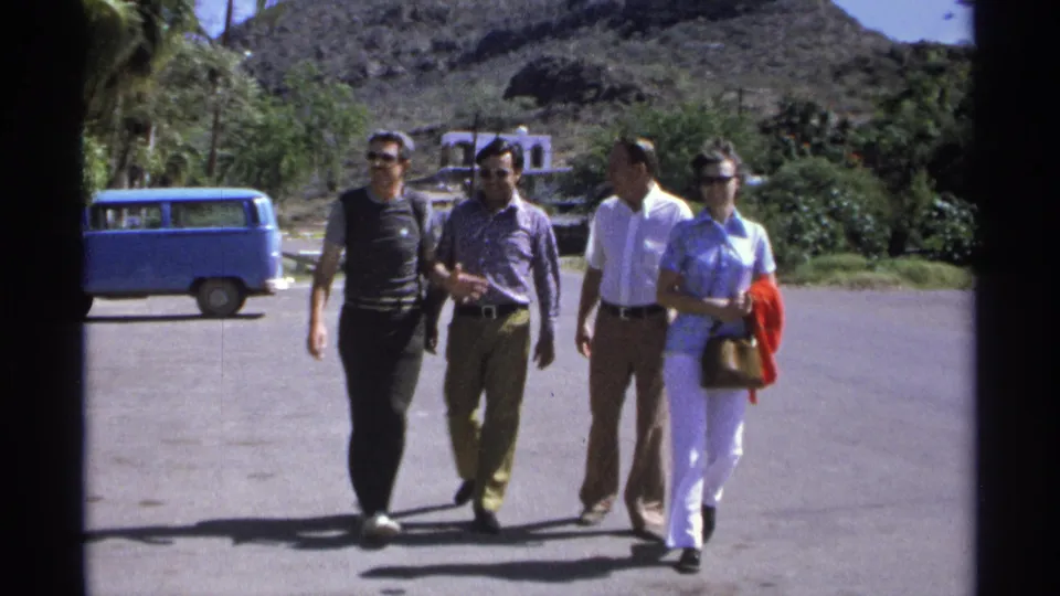 Four adults continue walking toward the camera in Guaymas, Mexico, wearing sunglasses and casual 1970s clothing near a blue van.