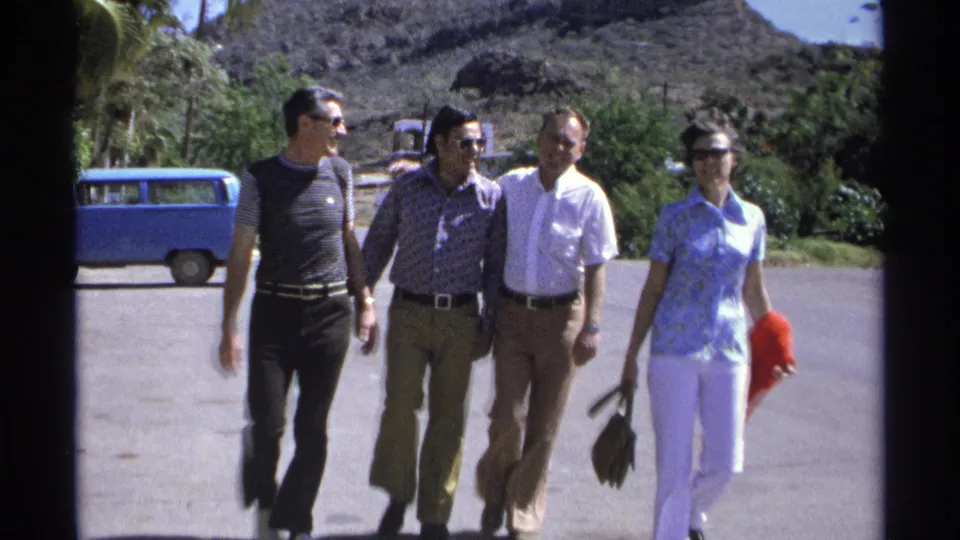 In the 1975 Guaymas home movie, four adults walk side by side toward the camera, with sunlit pavement, palms, and hillside visible.