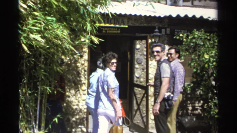 Adults on a 1975 Guaymas vacation gather at an entrance as two men in sunglasses glance back at the home-movie camera.