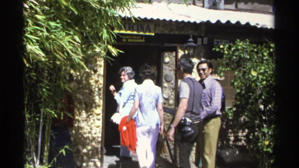 In this 1975 Guaymas home-movie frame, adults enter a stone-fronted establishment while one woman smiles toward the camera.