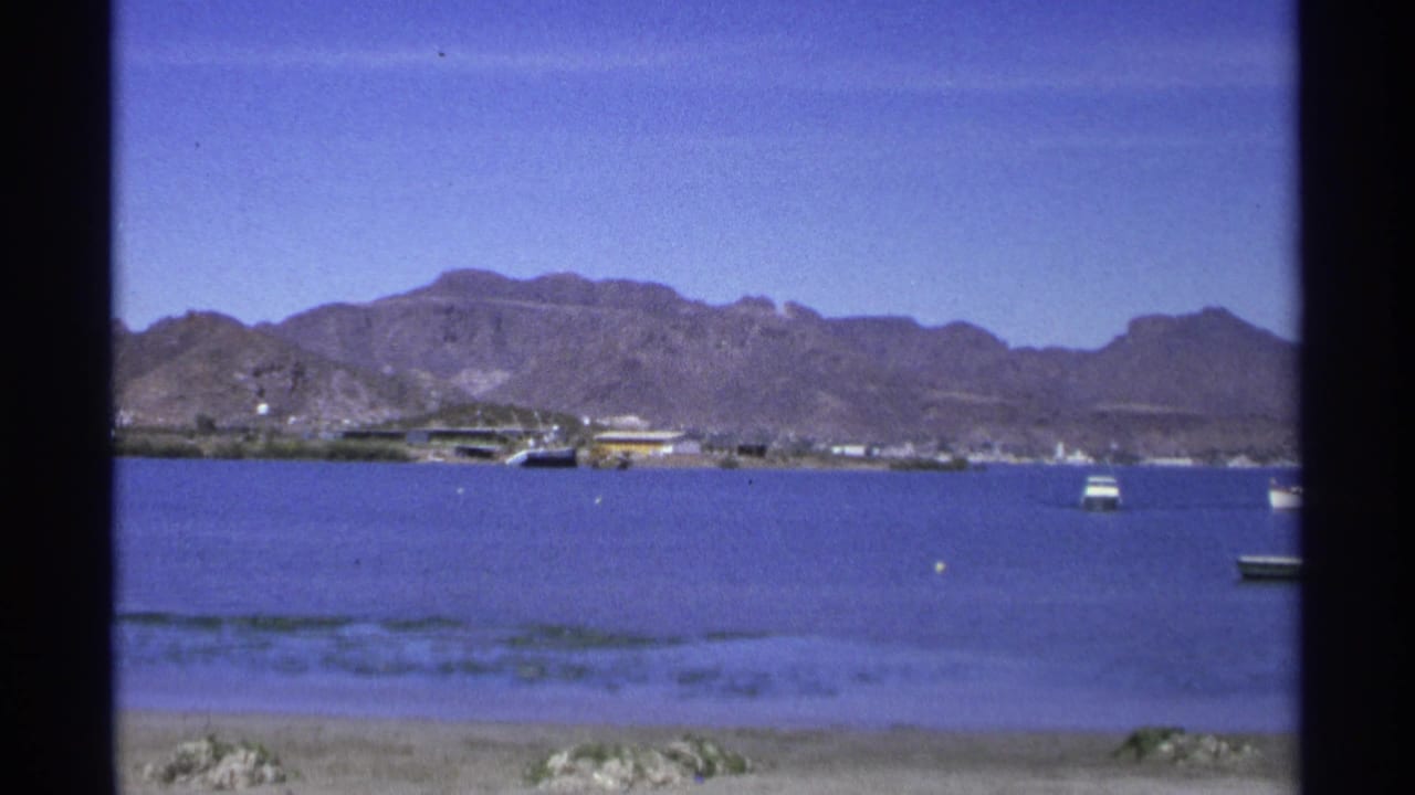 Calm harbor water with small boats, shoreline buildings and mountains beyond.