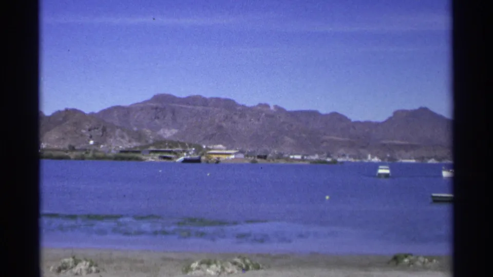 1975 Guaymas harbor view with blue water, anchored boats, distant shoreline structures and rugged mountains under a clear sky.