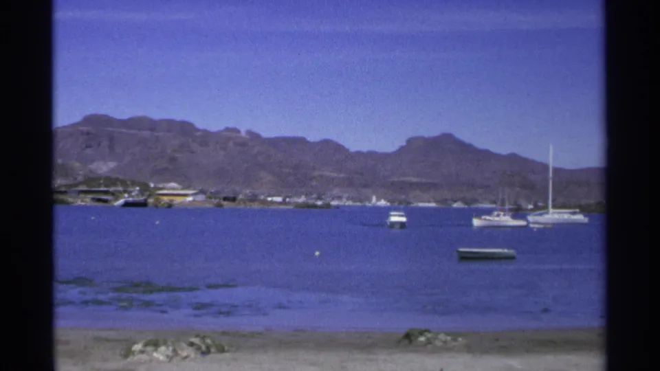 1975 Guaymas harbor frame with sailboats and a small motorboat on blue water, backed by shoreline structures and mountains.