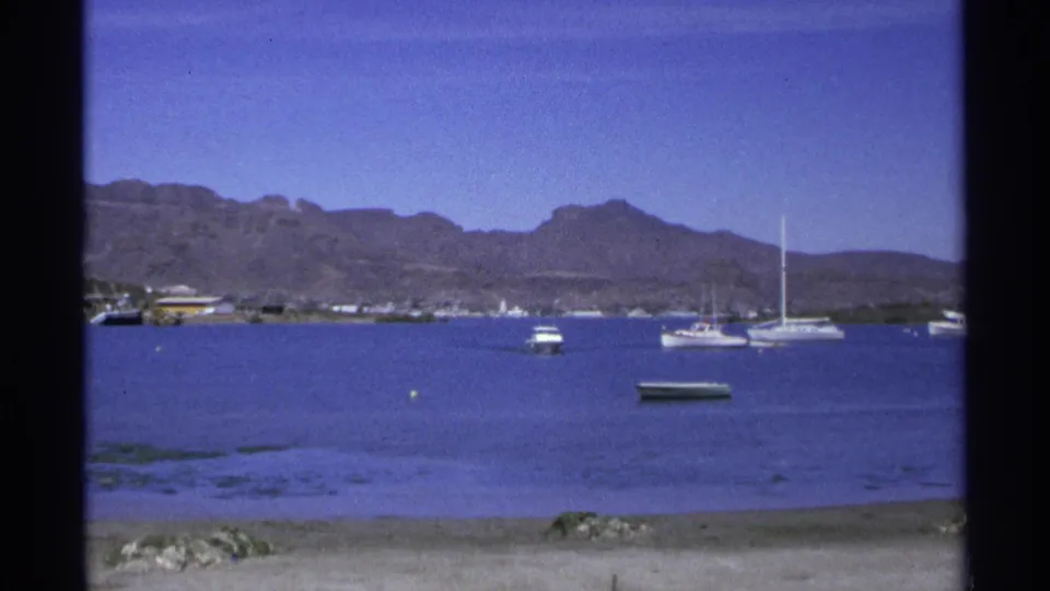 1975 Guaymas waterfront view with several anchored boats, visible sailboat masts, calm water and a rugged mountain backdrop.