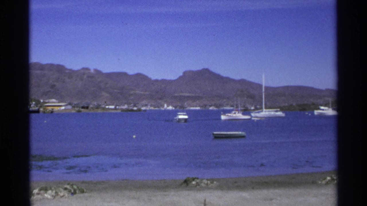 Blue bay with small boats, sailboats, sandy shore, and mountains in the background.
