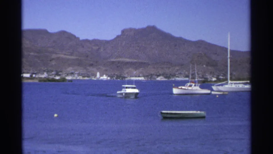 1975 Guaymas frame showing a white motorboat moving across calm bay water near anchored sailboats and a green rowboat.