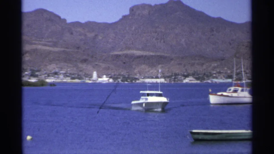 1975 Guaymas home-movie frame with a white motorboat closer to shore, a sailboat nearby, and mountains behind the waterfront.