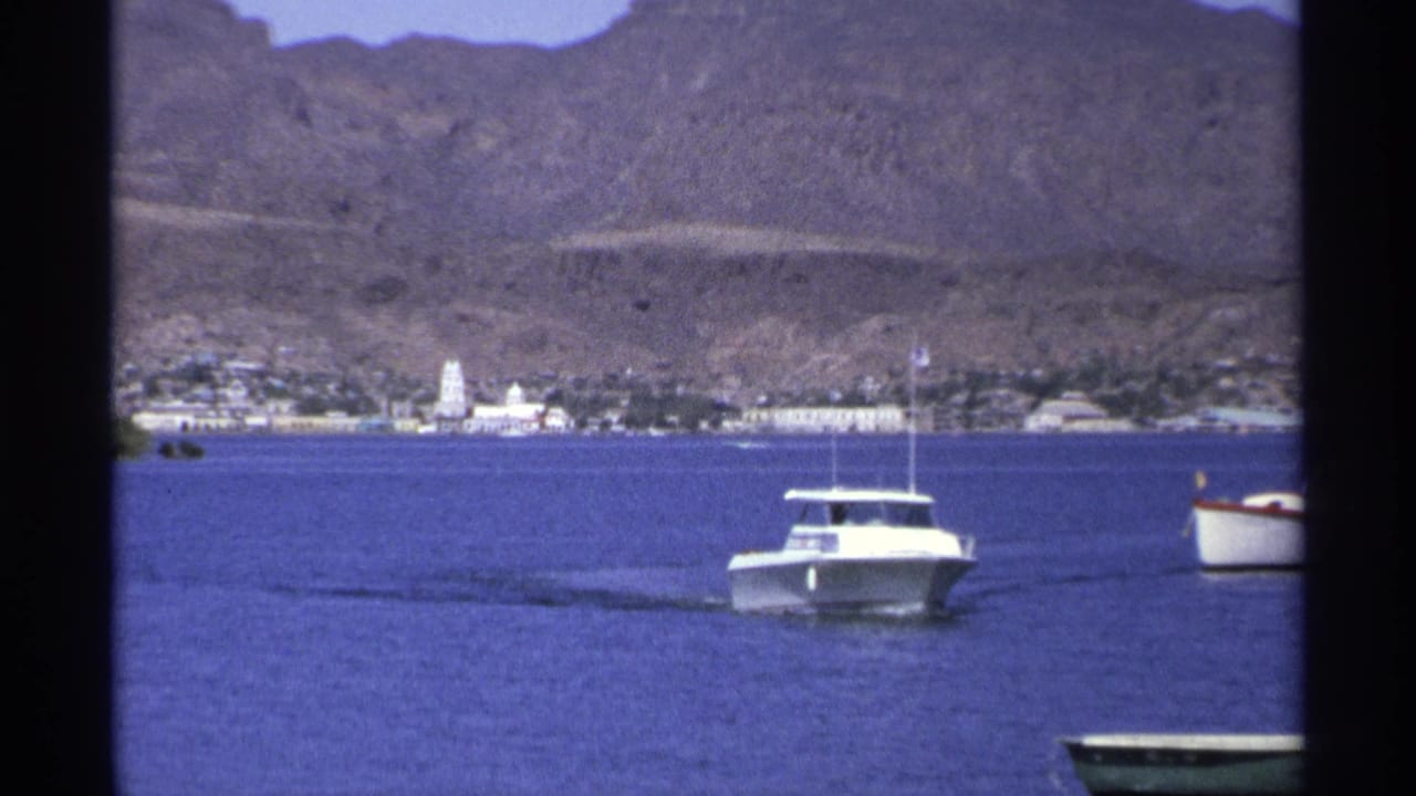 White motorboat on blue harbor water with a coastal town and dry mountains behind it.