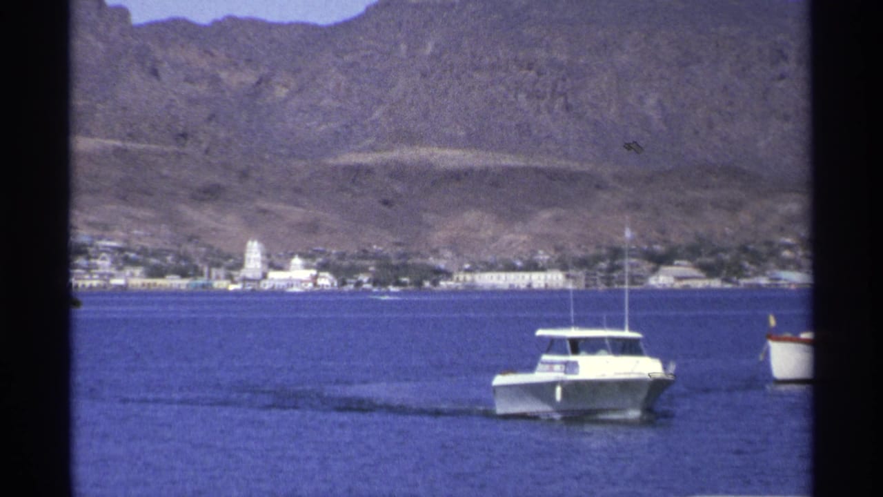 White motorboat farther right on calm water with shoreline buildings and mountains behind.