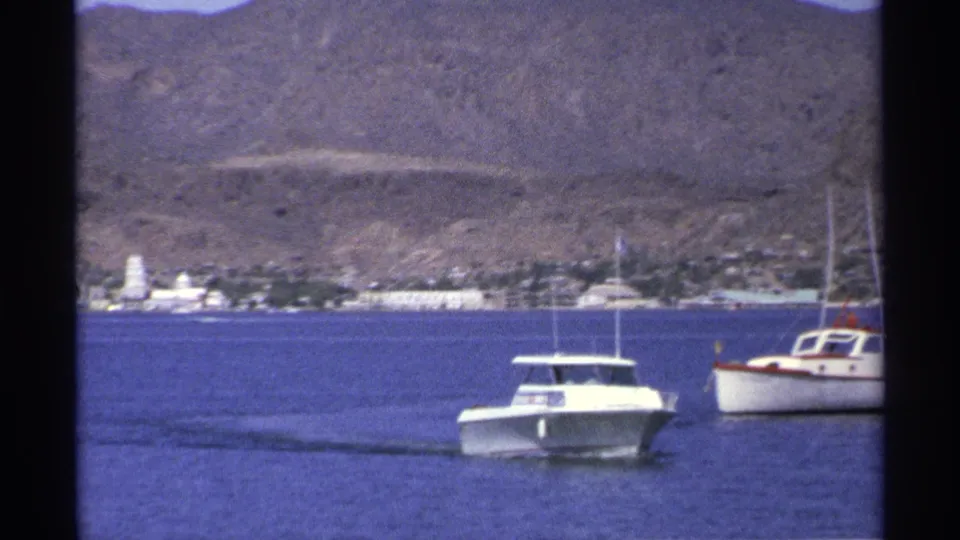 1975 Guaymas home-movie frame showing a white motorboat near another small boat, with town buildings below rugged hills.