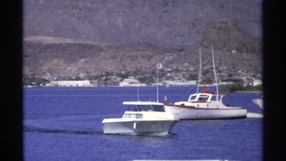 1975 Guaymas frame with a white motorboat passing a small mast boat on calm water, with shoreline buildings and dry hills beyond.