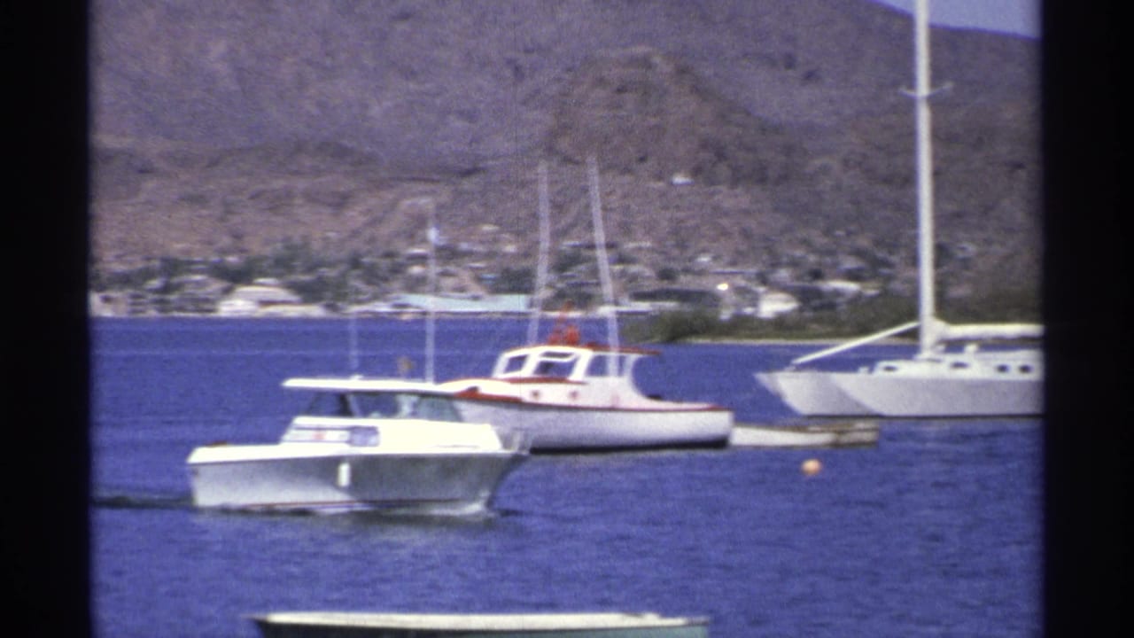 1975 Guaymas harbor view with a white motorboat in front of sailboats on deep blue water and arid hills in the background.