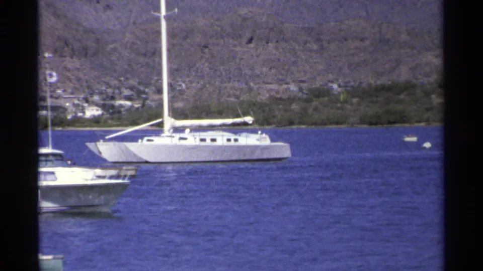 A large white sailboat or catamaran crosses the Guaymas harbor frame, with a smaller boat edge and hillside shore visible.