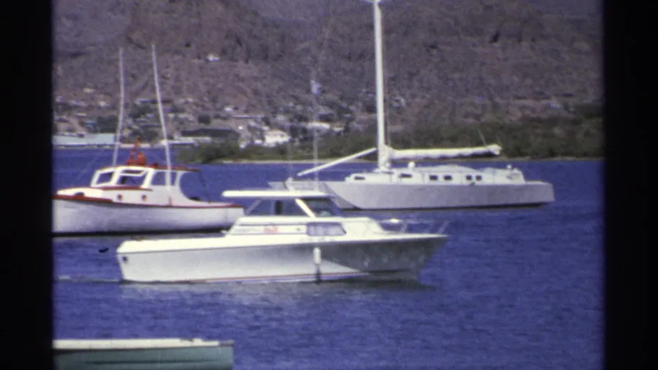 A white cabin motorboat moves across the foreground in Guaymas harbor, with sailboats, buoys, shoreline buildings, and dry hills beyond.