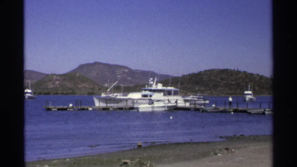 A 1975 home-movie frame shows a white yacht at a marina dock in Guaymas, with blue water, shoreline, and hills behind.