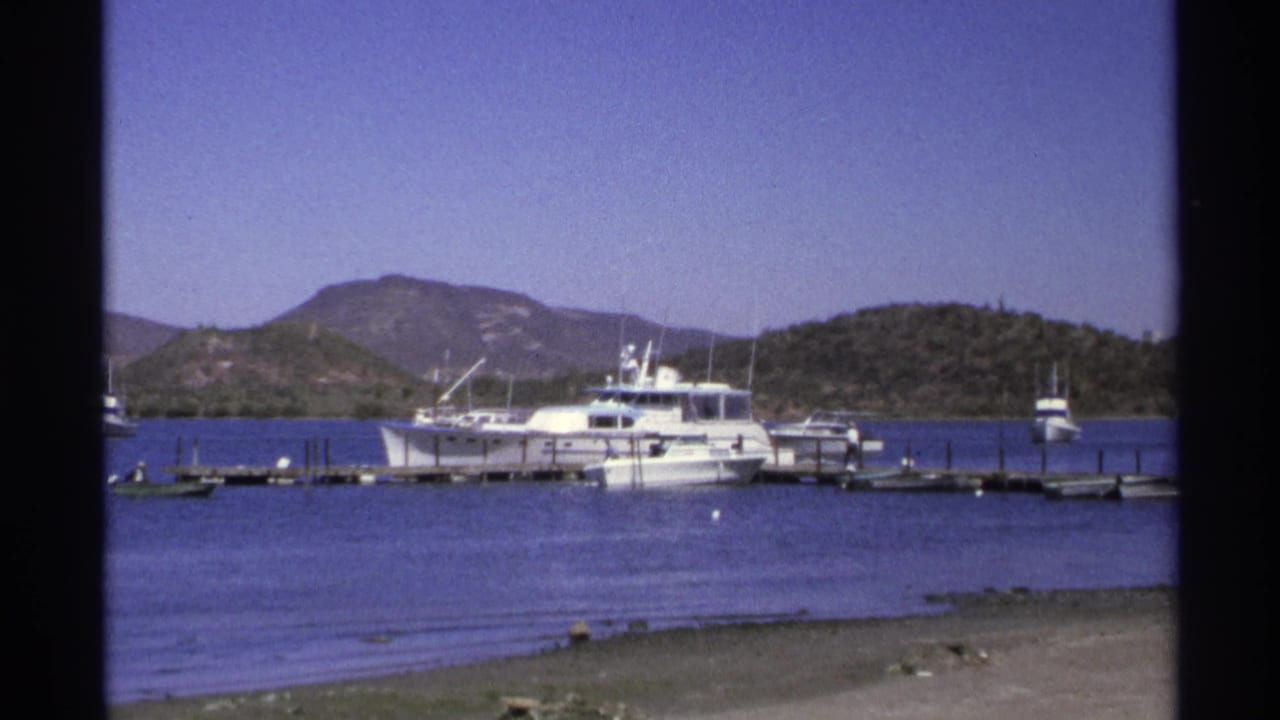 White boats and docks on a marina, seen across water with hills in the distance.