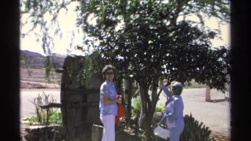 Guaymas home-movie frame from 1975 showing two women beside a leafy garden tree, with a road and low hills visible beyond.