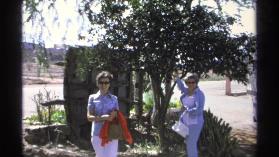 Two women in sunglasses appear outdoors in Guaymas, Mexico, with one holding a red scarf and the other reaching toward the tree canopy.