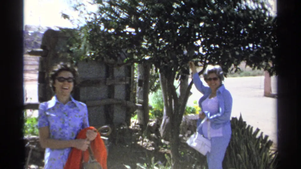 A closer 1975 home-movie frame shows a smiling woman holding a red scarf while another woman touches leaves near a roadside tree.