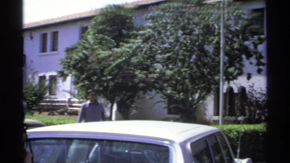 1975 Guaymas home-movie frame showing a white hotel exterior, trees, parked car roof, and an adult man near the walkway.