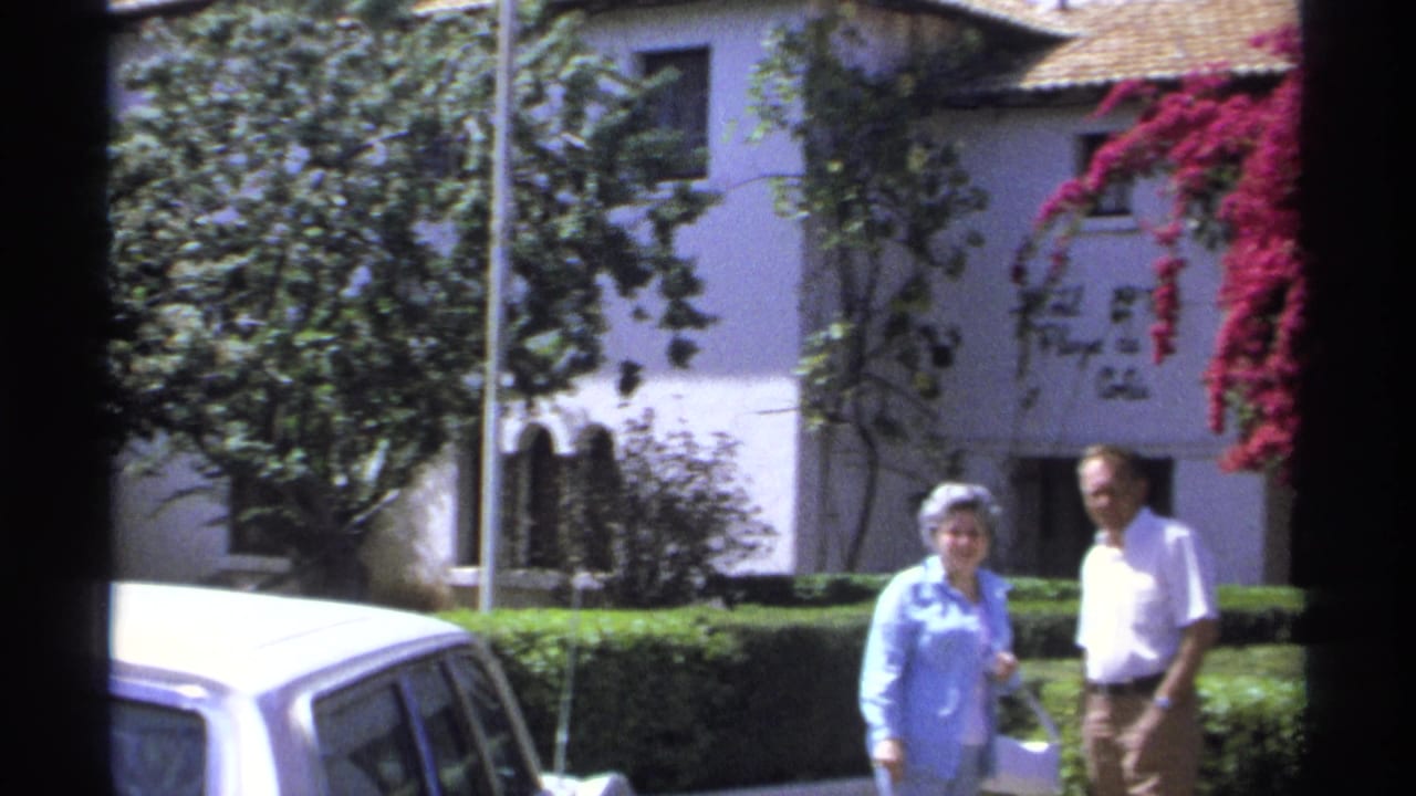 Older couple walking beside hedges near a white hotel and bright pink bougainvillea.