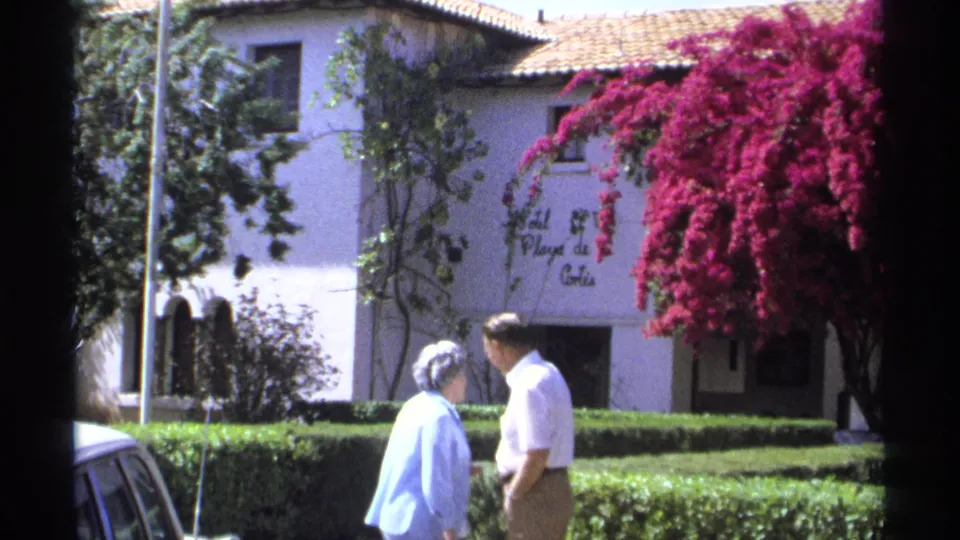 1975 Guaymas home-movie frame showing a couple conversing near the Hotel Playa de Cortés sign, hedges, and bougainvillea.