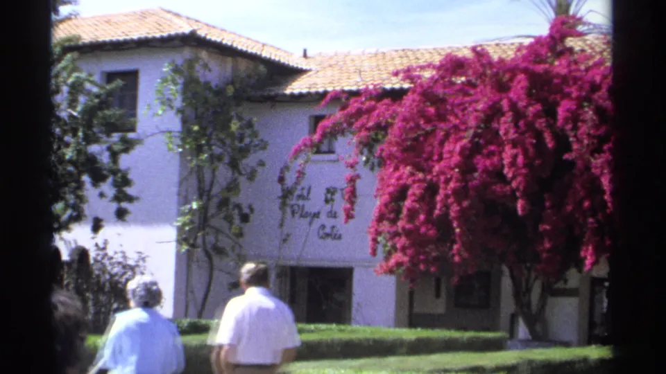 1975 Guaymas frame showing the couple walking away near the Hotel Playa de Cortés entrance, with hedges and pink bougainvillea.