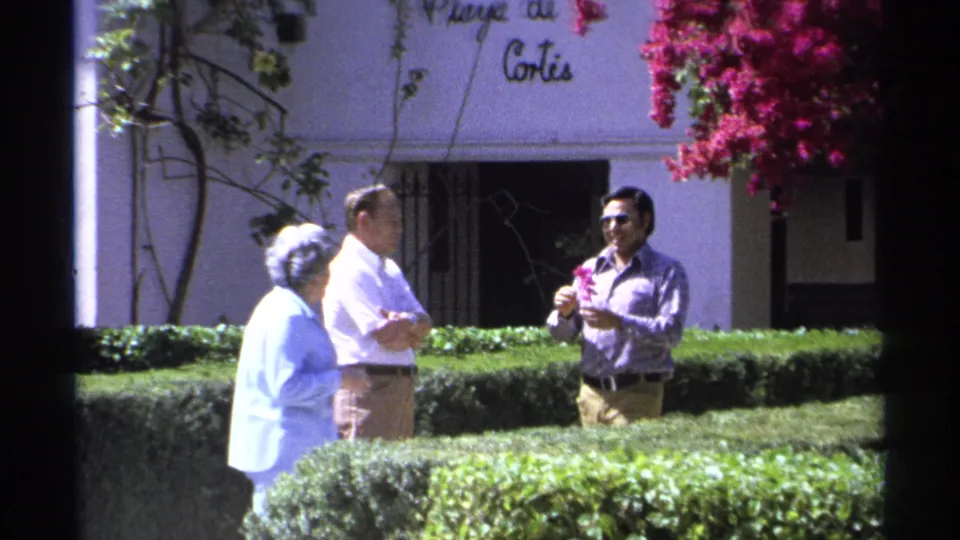 In the 1975 Guaymas garden scene, a man holding a red flower faces two adults near partial Playa de Cortés signage.