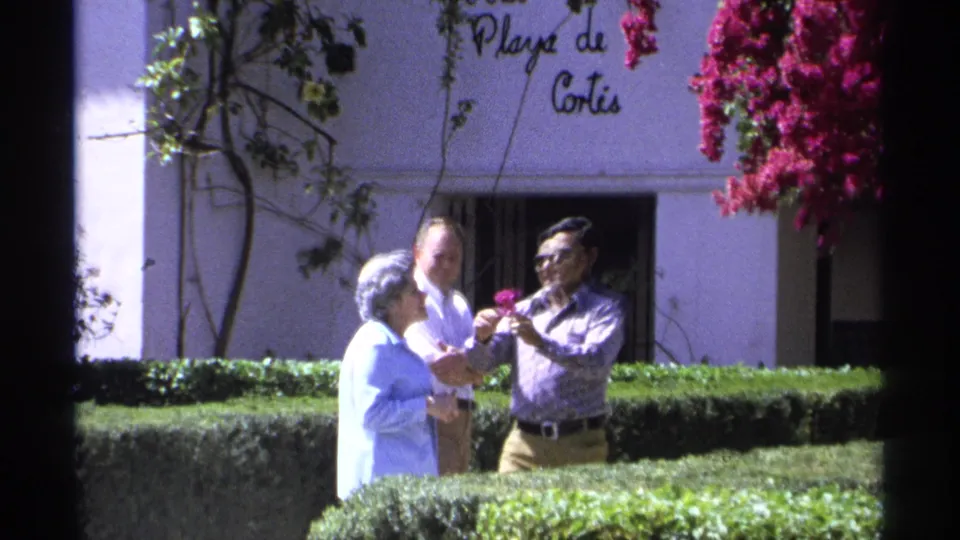 1975 Guaymas home-movie frame with a man in sunglasses placing a pink flower in an elderly woman's gray hair near a Playa de Cortes wall.