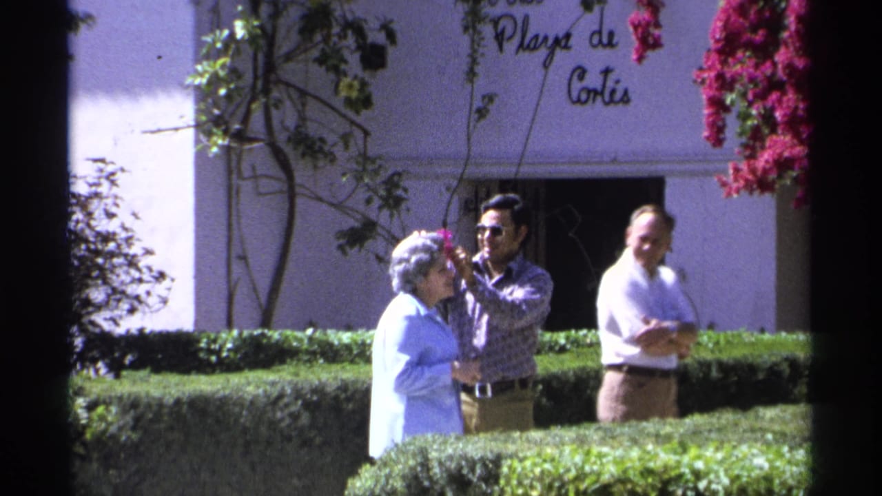 Three adults stand near hedges as a man adjusts a flower in an elderly woman's hair.