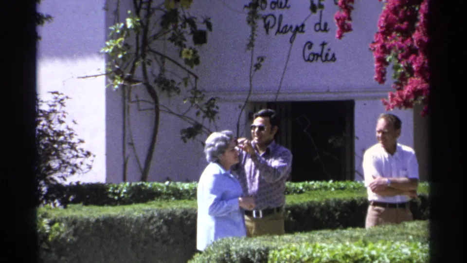 1975 Guaymas footage shows the flower being positioned in an elderly woman's hair, with hedges and the Playa de Cortes wall behind them.